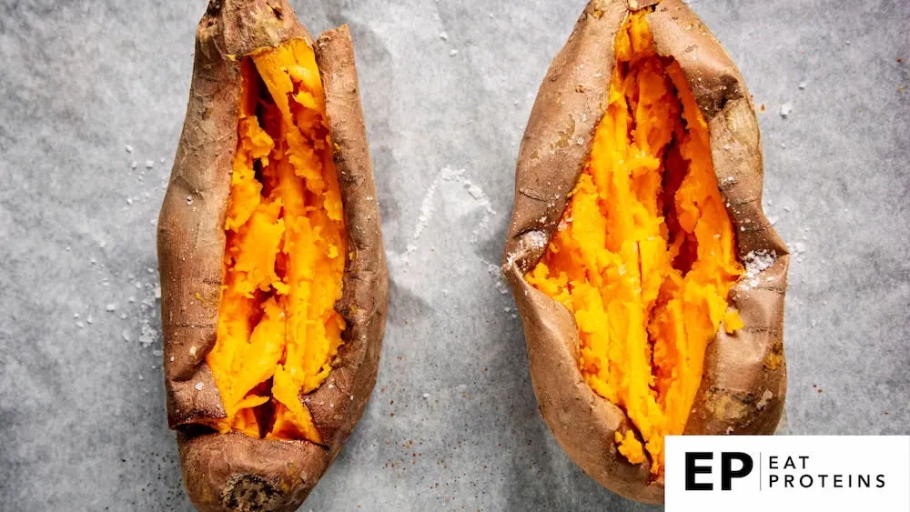 A rustic kitchen counter with a pile of baked sweet potatoes, surrounded by ingredients like herbs, olive oil, and spices