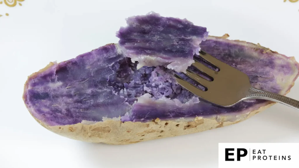 The image shows a baked "Beni Imo," a type of Okinawan purple sweet potato. The potato has been cut open, revealing its vibrant purple flesh. A fork is lifting a piece of the soft, mashed sweet potato, highlighting its smooth texture. The outer skin is light brown, contrasting with the rich purple interior.