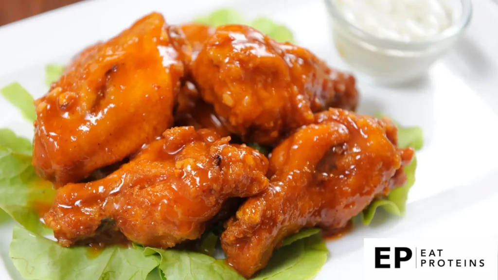 Close-up of saucy buffalo wings with a rich orange glaze, served on a bed of fresh lettuce, accompanied by a small bowl of creamy dipping sauce in the background