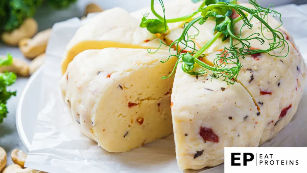 Close-up image of a round block of cashew cheese with herbs and sun-dried tomatoes. The cheese is sliced open, revealing a smooth, firm texture. It is garnished with fresh pea shoots and placed on parchment paper. 