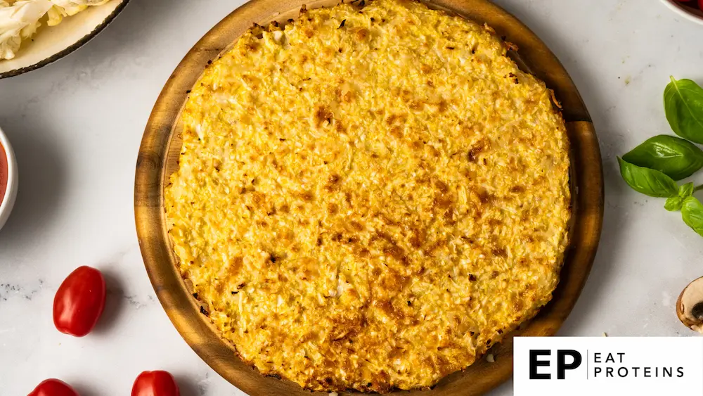 The image shows a large, golden-brown cauliflower breadstick or flatbread on a wooden board. The texture suggests a cheesy, crispy exterior, making it a keto-friendly, low-carb option. Surrounding the dish are fresh ingredients like cherry tomatoes, basil leaves, and mushrooms, hinting at potential toppings or accompaniments