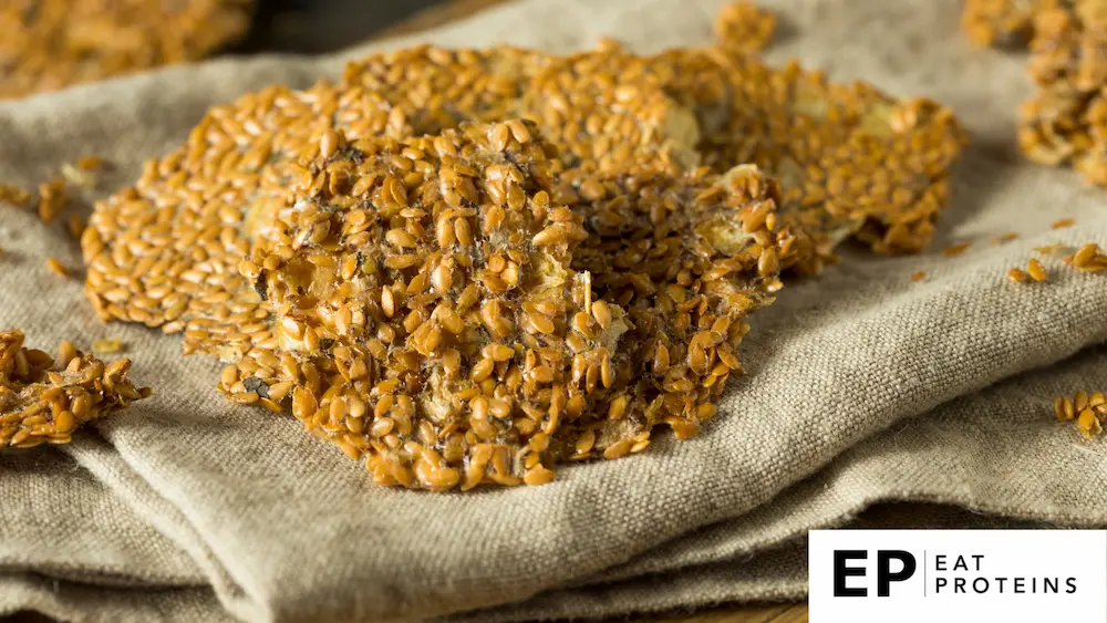 Close-up image of dehydrated flaxseed crackers on a folded beige cloth. The crackers have a crispy texture, made from whole flaxseeds that are compacted together, with some seeds scattered around. Their golden-brown color contrasts with the natural fabric beneath.