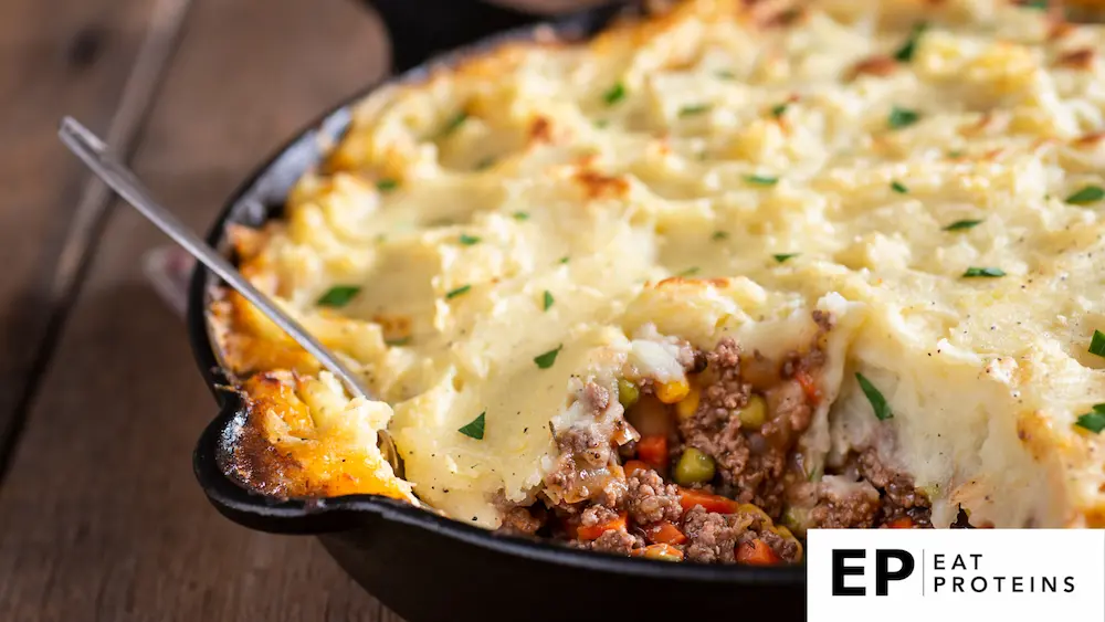 The image features a hearty shepherd's pie served in a cast-iron skillet. The dish has a golden, creamy mashed potato topping, sprinkled with fresh herbs for a hint of color. The filling underneath reveals a mixture of ground meat or a plant-based protein alternative, mixed with a variety of vegetables such as peas, corn, carrots, and onions, creating a vibrant and inviting combination