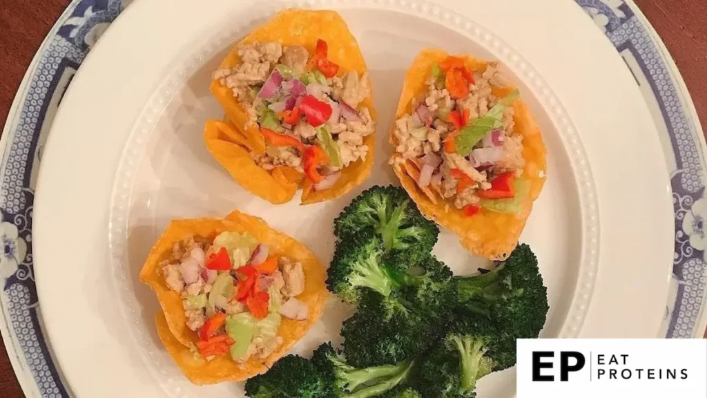 The image shows three low-carb taco cups made from a crispy cheese shell, filled with a mix of ground meat, diced red onions, lettuce, and red bell peppers. The taco cups are placed on a white plate with a side of steamed broccoli, making it a balanced, keto-friendly meal