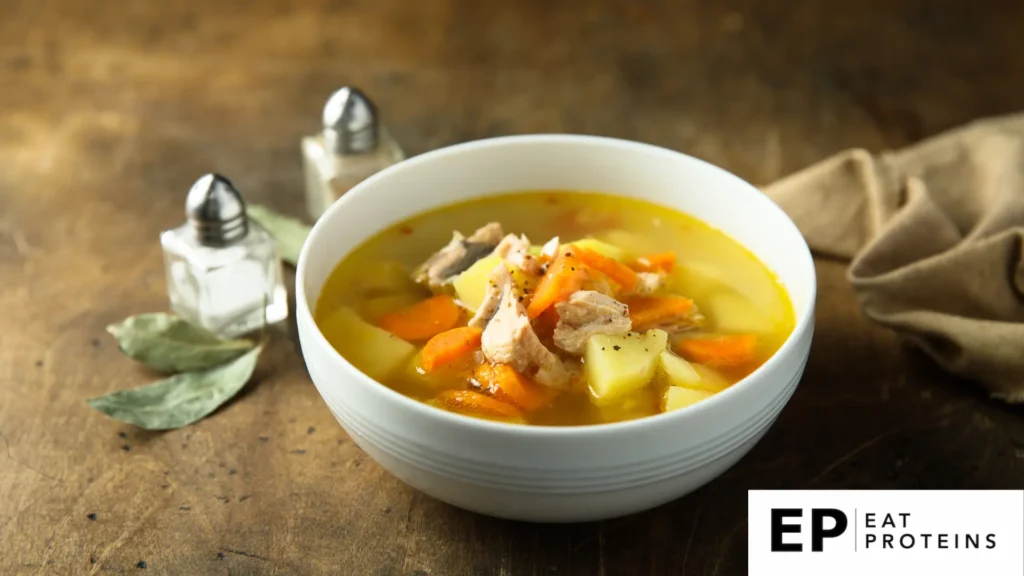 A rustic kitchen table with a steaming bowl of Mediterranean fish soup surrounded by fresh herbs, tomatoes, and a loaf of crusty bread