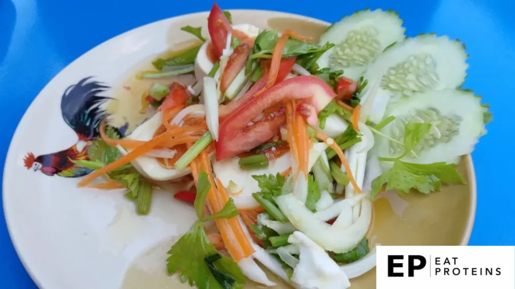 The image shows a plate of "Munata," a fresh Okinawan vegetable salad. The dish is made with sliced tomatoes, cucumbers, onions, carrots, and herbs like cilantro, arranged neatly on the plate. The vegetables are vibrant and crisp, served on a plate with a rooster design.