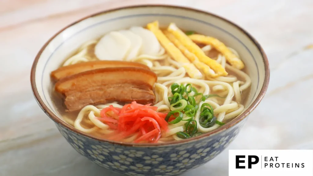 The image shows a bowl of Okinawa soba, a traditional Okinawan noodle soup. The dish includes thick noodles in a clear broth, topped with slices of braised pork belly, green onions, kamaboko (fish cake), thin strips of egg, and pickled ginger