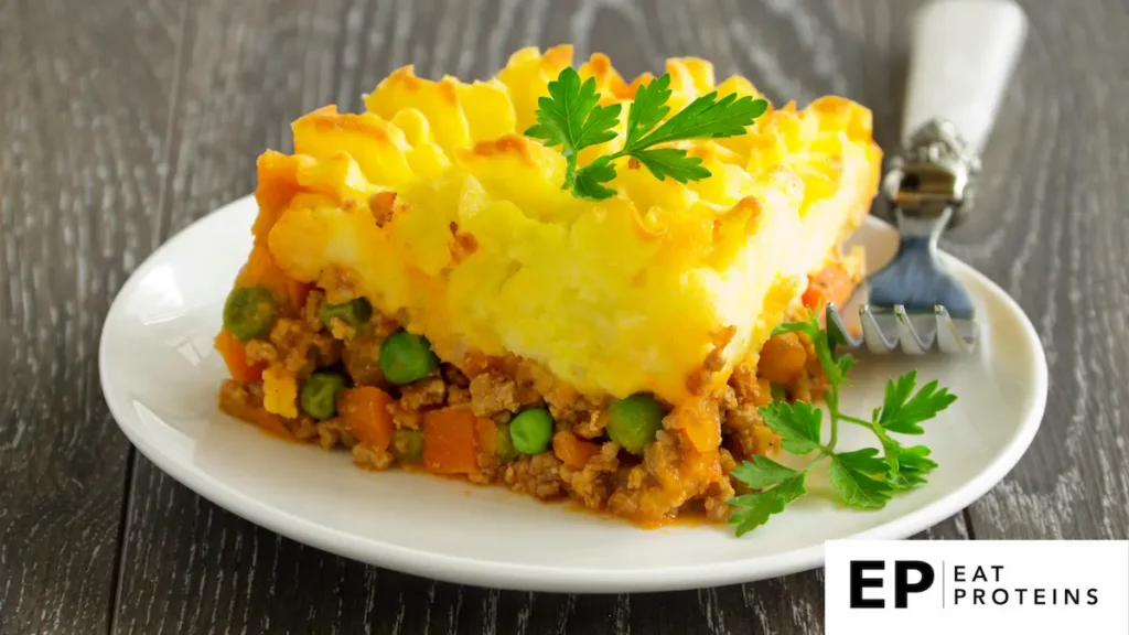 The image depicts a slice of shepherd's pie, a classic comfort dish. The top layer consists of fluffy, golden-brown mashed potatoes, piped decoratively and baked until slightly crispy. Below, there is a savory filling made of ground meat (typically lamb or beef), mixed with vegetables such as peas and carrots, all coated in a rich sauce. The dish is garnished with a sprig of fresh parsley, adding a touch of color