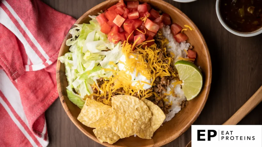 The image shows a bowl of "Taco Rice," a popular Okinawan dish. It consists of seasoned ground beef over rice, topped with shredded lettuce, diced tomatoes, grated cheese, sour cream, and a lime wedge. Tortilla chips are placed on the side, and a red-and-white napkin is visible next to the bowl