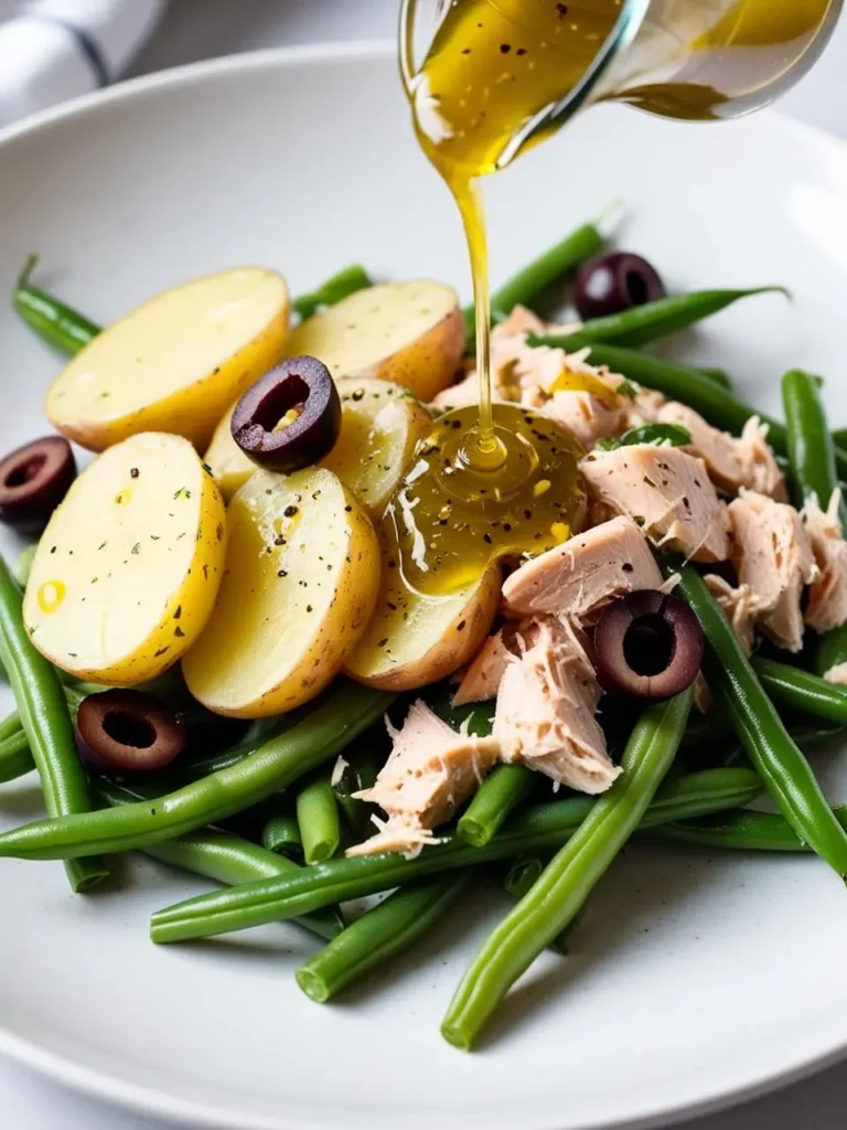 a salad with green beans, potatoes, tuna, and olives on a white plate. a clear glass pitcher is pouring a yellow dressing over the salad. the potatoes are sliced and yellow. the tuna is flaked and light pink. the olives are dark and round. the green beans are long and green