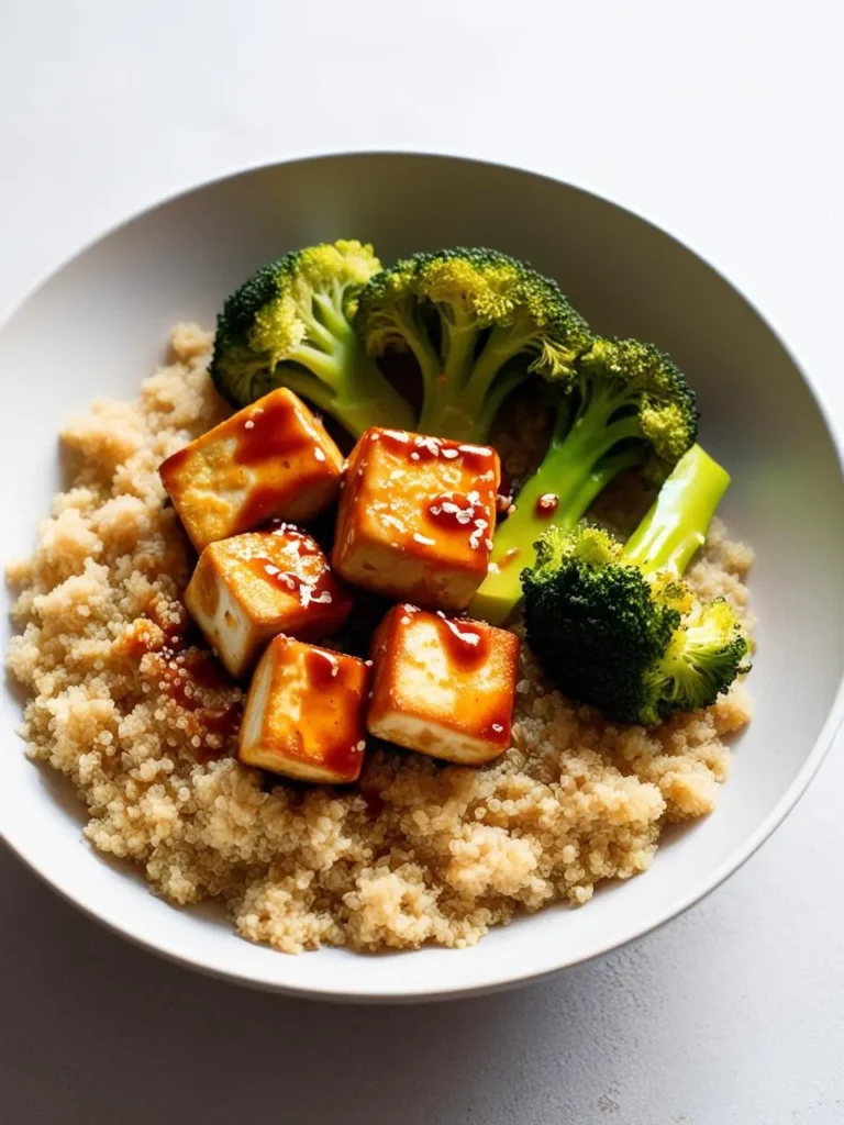 a white bowl filled with quinoa, topped with golden-brown tofu cubes drizzled with sauce and sesame seeds. vibrant green broccoli florets are arranged beside the tofu. the dish is presented simply, showcasing the textures and colors of the ingredients. the background is a neutral, light surface